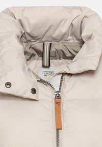 Helle beige Daunenjacke mit hohem Kragen, Reißverschluss mit orangefarbenem Ziehband, metallischen Druckknöpfen und kontrastierender Innenauskleidung mit Streifen.