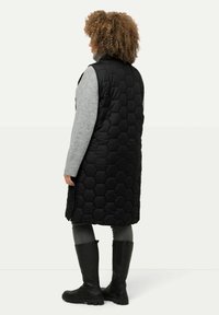 Schwarze, hexagonale gepolsterte Weste, die bis zur Mitte des Oberschenkels reicht. Getragen über einem grauen Langarmoberteil, kombiniert mit dunklen Jeans und knielangen schwarzen Stiefeln.