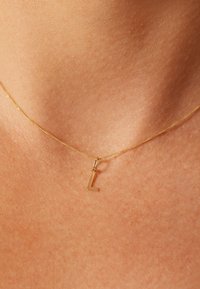 Collier en or avec un pendentif en forme de "L" minuscule, texture lisse et design minimaliste, posé sur une peau légèrement bronzée.