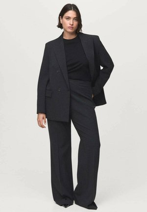 Žena s krátkými tmavými vlasy, oblečená v oversized blejzru s tmavým proužkem, odpovídajících širokých kalhotách a černém topu, stojící na jednobarevném pozadí.