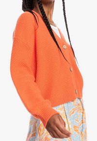 Helles, oranges, kurzes Strickjacken-Cardigan mit strukturierter Strickweise, ausgestattet mit drei großen Holzknoepfen und gerippten Buendigungen, kombiniert mit einem Rock mit Blumenmuster.