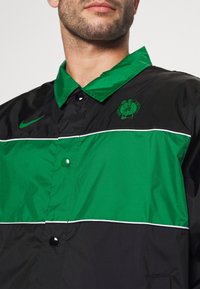 Zwarte en groene windbreaker met een knoopsluiting, contrasterende strepen en het Boston Celtics-logo op de linkerborst. Glad, glanzend oppervlak.