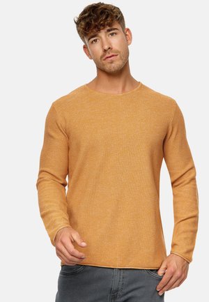 Ung mand med kort krøllet hår iført en tekstureret langærmet orange sweater og grå bukser mod en ensfarvet baggrund.