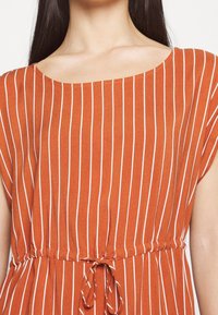 Rostfärgad klänning med vita vertikala ränder. Har en bred halsringning, kortärmar och en samlad midja med ett knytband. Slät textur.