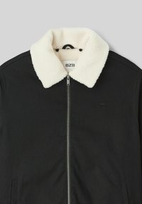 Veste noire avec un col en fausse fourrure blanche, fermeture éclair sur le devant et deux poches latérales. Présente une étiquette de marque discrète sur la poitrine gauche.