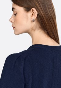 Junge Frau mit glattem braunem Haar, die silberne Creolen und einen marineblauen Pullover trägt, im Profil vor einem weißen Hintergrund abgebildet.