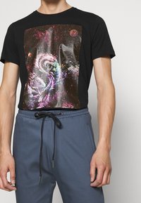 Svart kortärmad t-shirt med en färgglad kosmisk grafisk tryck. Matchas med blå byxor med elastisk midja, som har dragsnörning.