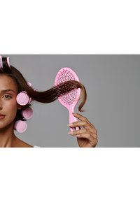 Brosse à cheveux en plastique de couleur rose clair, dotée d'une tête ovale perforée et d'un manche droit, tenue dans une main avec des cheveux stylés et des bigoudis.