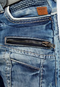 Jeans in denim blu con una tasca con zip, cuciture a contrasto e un'etichetta in pelle. Il tessuto presenta leggere scoloriture e variazioni di texture.