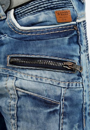 Jeans in denim blu con una tasca con zip, cuciture a contrasto e un'etichetta in pelle. Il tessuto presenta leggere scoloriture e variazioni di texture.