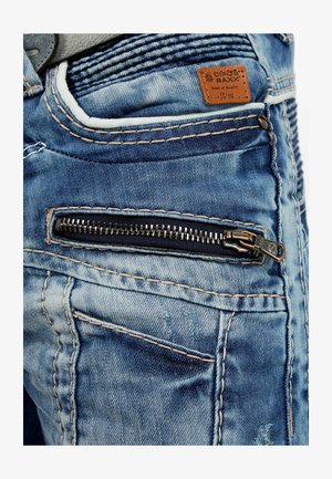 Blue denim jeans with a zippered pocket, contrasting stitching, and a leather patch label. The fabric shows slight fading and texture variations.