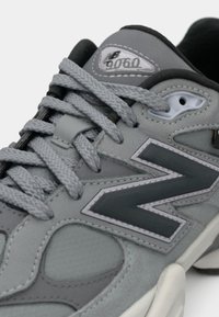 Graue New Balance 9060 Sneakers mit dicken grauen Schnürsenkeln, schwarzen und hellgrauen Schichten und einem großen schwarzen "N"-Logo an der Seite.