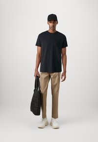 Schwarzes Baumwoll-T-Shirt, beigefarbene, maßgeschneiderte Hose, weiße Sneakers, hält eine schwarze gesteppte Tragetasche, einfarbiger Hintergrund.