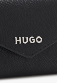 Portafoglio in pelle nera con finitura testurizzata, caratterizzato da un logo argentato "HUGO" sul davanti e un design a pattina diagonale.