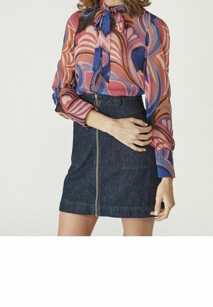 Donna che indossa una blusa con fantasia multicolore e collo a fiocco e una gonna in denim scuro con chiusura a cerniera frontale.