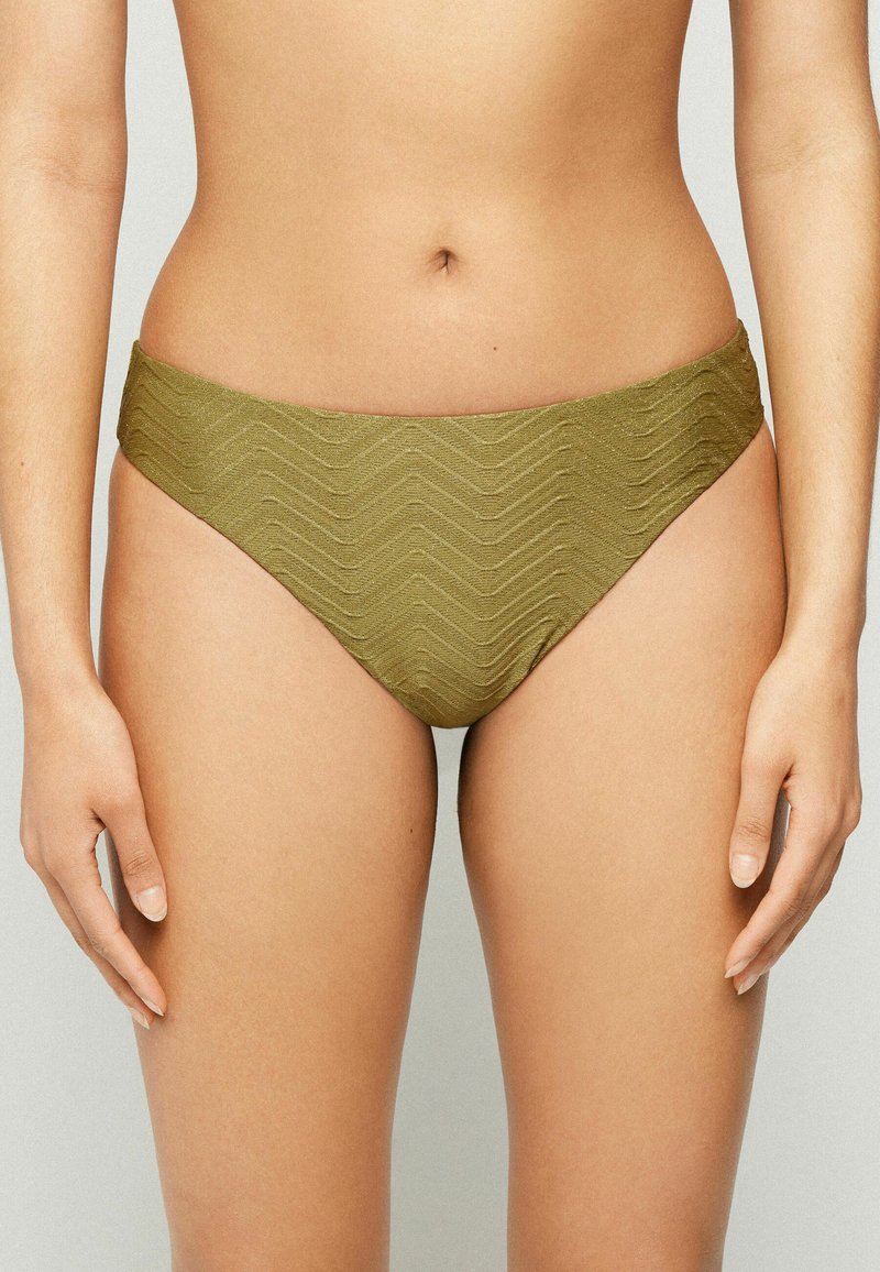 Femme portant un bas de bikini vert olive avec un motif en zigzag texturé, montrée de la taille aux cuisses supérieures sur un fond neutre.