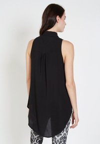 Mujer vista desde atrás, vestida con una túnica negra sin mangas con un cuello alto y pantalones a rayas en blanco y negro, sobre un fondo blanco.