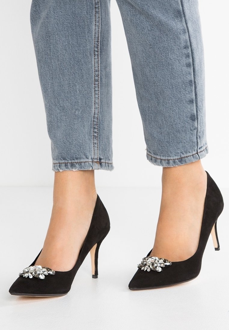 Escarpins en daim noir à talons hauts avec pointes pointues et strass décoratifs, portés avec un jean bleu clair raccourci sur un sol blanc.