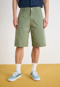 Green cotton shorts with a straight fit, solid texture, front pockets, and a button closure; paired with light blue sneakers and white socks.