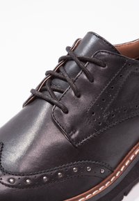 Chaussure habillée à bout fleuri en cuir noir avec perforations brogue, lacets noirs ronds et semelle superposée avec des accents de couture marron et blanche.