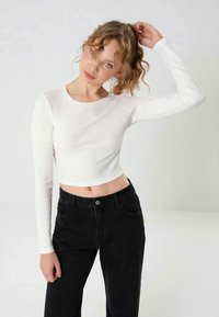 Camisa branca cropped de manga longa com decote redondo, combinada com jeans pretos de cintura alta, com bolsos e fecho de botão à frente.
