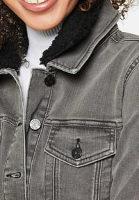 Graue Denimjacke mit schwarzem Fleece-Kragen. Verfügt über zwei schwarze Knöpfe, einen silbernen Logo-Knopf und Details an der vorderen Brusttasche.