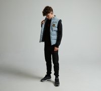 Gilet matelassé bleu clair avec des accents noirs, fermeture éclair et écusson de logo sur une chemise à manches longues noire et un pantalon de jogging assorti. Baskets noires.