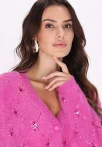 Rosa, flauschige Strickjacke mit Pailletten, runder Knopfverschluss, tiefem V-Ausschnitt und langen Ärmeln. Das Model posiert mit der Hand am Kinn und trägt auffällige Ohrringe.