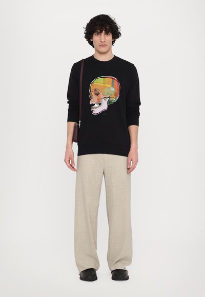 Sort sweatshirts med et farverigt kraniumgrafik, parret med lyse beige bukser og sorte sko. Modellen står med en brun skuldertaske.