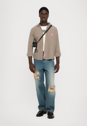 Geruite shirt met rode patch, wit t-shirt, versleten lichtblauwe spijkerbroek en zwarte schoenen. Kleine zwarte tas over de schouder.