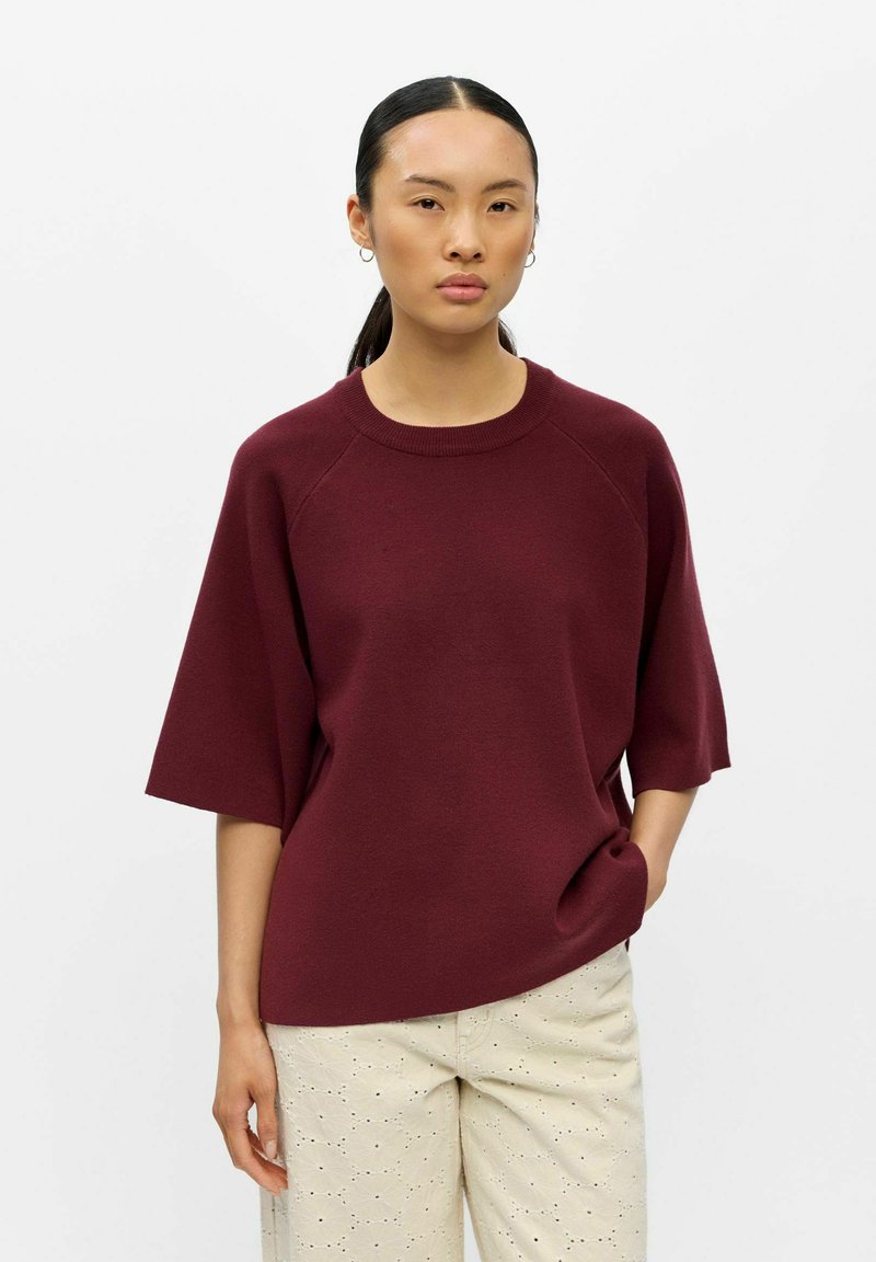 Femme aux cheveux foncés attachés, portant un pull bordeaux à manches courtes et un pantalon beige clair à motif œillet, debout devant un fond blanc.