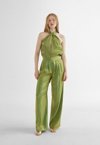 Conjunto de satén verde que presenta una blusa de cuello halter con detalle de ojal y pantalones de pierna ancha. Tejido texturizado con un ligero brillo.