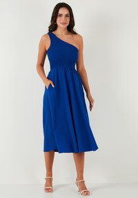 Blue one-shoulder dress with a smocked bodice, flowing midi skirt, and side pockets. Made of lightweight fabric with a textured finish.