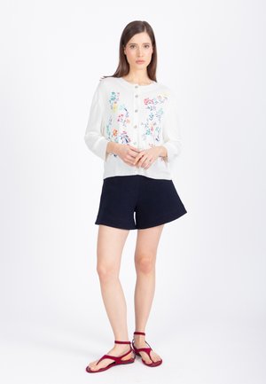 Žena stojaca, oblečená v bielom kvetovanom zapínacom kardigáne, námorníckych šortkách a červených sandáloch s remienkom okolo členka na bielom pozadí.