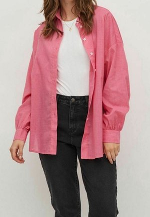 Kobieta nosząca luźną różową koszulę na guziki nałożoną na biały top, w połączeniu z czarnymi dżinsami z wysokim stanem, stojąca na jednolitym tle.