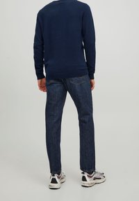 Mężczyzna ubrany w ciemnoniebieski sweter, ciemnoniebieskie dżinsy i białe trampki, stojący tyłem na jednolitym tle.