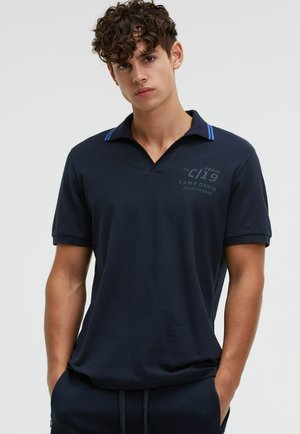 Junger Mann mit lockigem Haar, der ein marineblaues Polohemd mit blauen Streifen am Kragen und Text auf der Brust trägt.