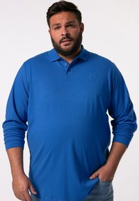 Modrá polokošile s dlouhými rukávy vyrobená z měkké látky, s límečkem a decentním logem na hrudi, doplněná džínovými kalhotami.