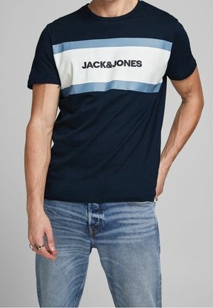 Mann trägt marineblaues T-Shirt mit weißen und hellblauen horizontalen Streifen und dem Schriftzug "JACK & JONES", kombiniert mit hellblauen Jeans.