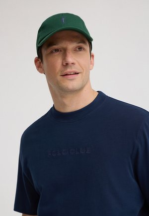 Man draagt een groene pet en een donkerblauw T-shirt met "POLO CLUB" in reliëf op de borst, kijkt lichtjes opzij.