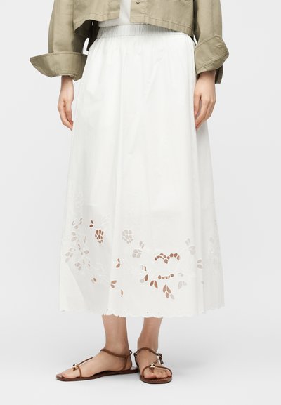 Γυναίκα που φοράει λευκή midi φούστα με κεντήματα με λουλουδάτα μοτίβα, σε συνδυασμό με καφέ επίπεδα σανδάλια και κοντό μπεζ μπουφάν.