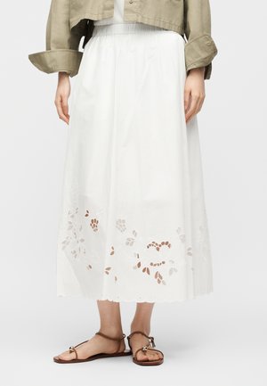 Kvinne iført hvit midiskjørt med blomsterutskjæring broderi, kombinert med brune flate sandaler og cropped beige jakke.