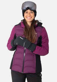 Lila und schwarze isolierte Skijacke mit Reißverschluss, kombiniert mit schwarzen Handschuhen und einer gestrickten Mütze. Das Model trägt Skibrille.