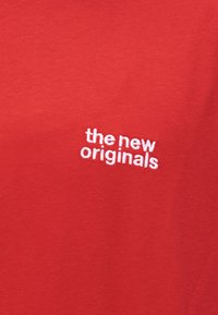 Crvena tkanina s bijelim vezom teksta "the new originals" blago nagnutog.