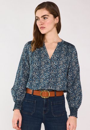 Femme portant un chemisier à manches longues bleu à fleurs, rentré dans un jean sombre, accessoirisé d'une ceinture marron avec une grande boucle dorée et des boucles d'oreilles créoles en or.
