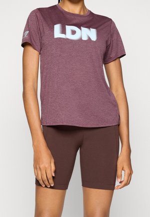 Femme portant un t-shirt bordeaux à manches courtes avec "LDN" en lettres blanches et un short marron foncé ajusté mi-cuisse sur un fond uni.