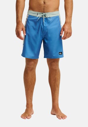 Man draagt blauwe boardshorts met lichtgrijze tailleband en trekkoord, staat blootsvoets tegen een effen achtergrond.