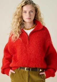 Junge Frau mit welligem blonden Haar, die einen roten, flauschigen Reißverschluss-Pullover, eine bernsteinfarbene Perlenkette und olivgrüne Cordhosen mit einem braunen Gürtel trägt.