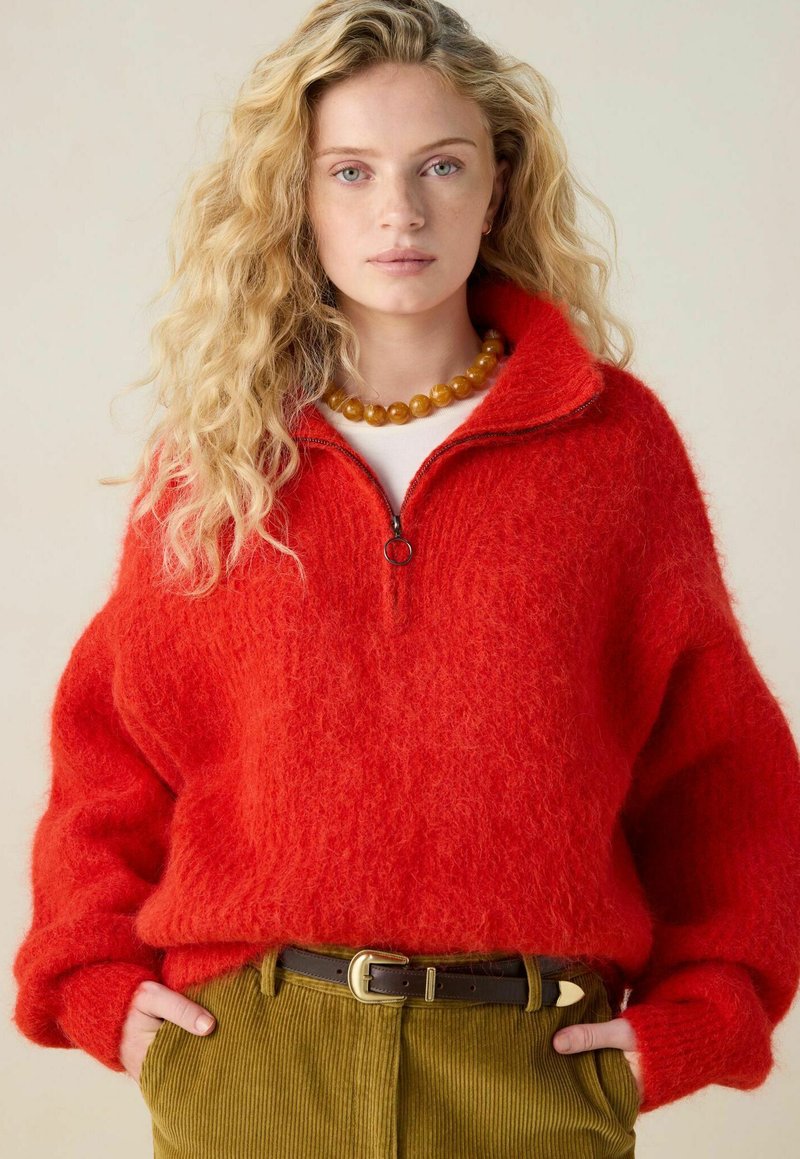 Junge Frau mit welligem blonden Haar, die einen roten, flauschigen Reißverschluss-Pullover, eine bernsteinfarbene Perlenkette und olivgrüne Cordhosen mit einem braunen Gürtel trägt.