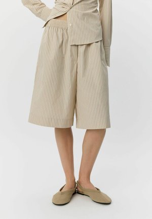 Luźne beżowo-białe szorty w prążki do kolan, noszone z pasującą koszulą na guziki i beżowymi, płaskimi butami z odkrytą piętą na pasku.